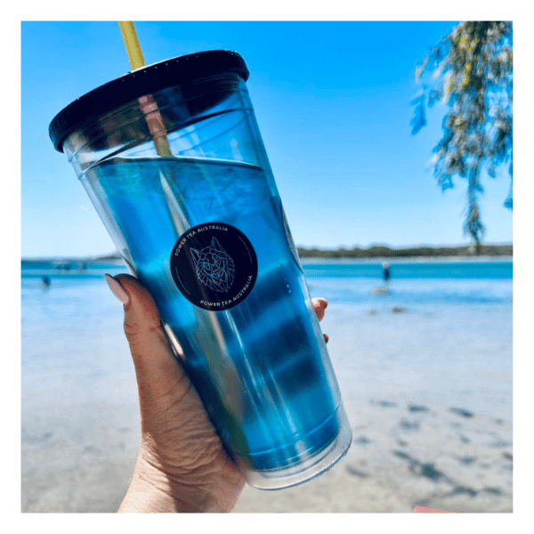 A blue Power Tea in a clear cup being held up to the camera with the beach in the back ground