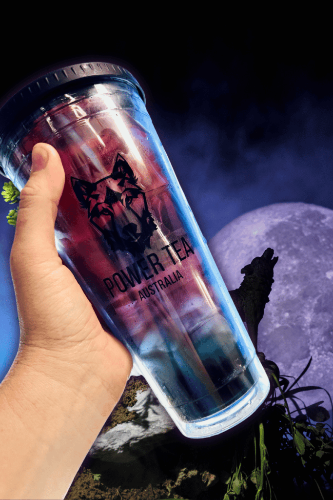 a hand holding a large cup of Annabelle Loaded Energy Power Tea. The beverage is deep red blending into a deep green in a clear cup, with a wolf howling at the moon in the back ground.